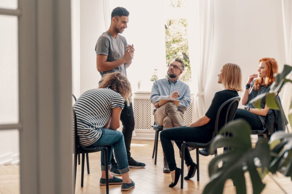 Young man standing & speaking with counselor and three others sitting.