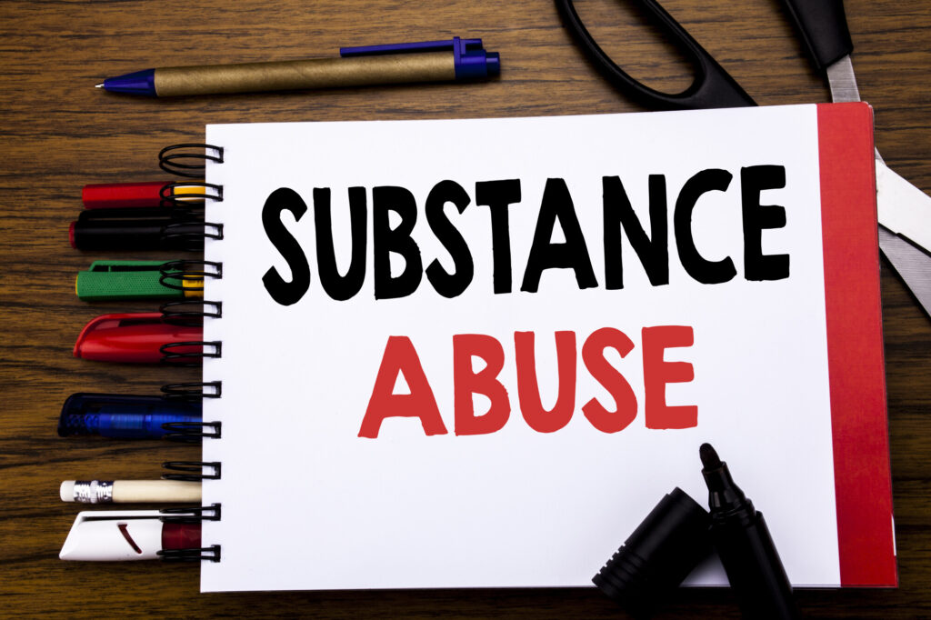 The word SUBSTANCE ABUSE written on a white notebook cover with a number of different color pens and a scissor behind the notebook