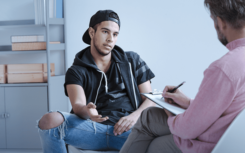 young man wearing jeans & black cap speaking with his counselor